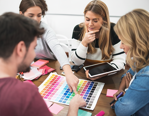 Un grupo de cuatro jóvenes —aparentemente estudiantes o profesionales del diseño— está reunido alrededor de una mesa de trabajo. En el centro, todos observan atentamente una gran paleta de colores impresa con múltiples tonos organizados en una cuadrícula. Uno de ellos, un joven de barba recortada, señala un color específico con el dedo mientras los demás siguen la selección con interés.
A la derecha, dos mujeres observan con atención: una con suéter claro y cabello rubio ondulado sujeta un bolígrafo mientras apoya el brazo sobre la mesa; la otra, con jersey blanco de rayas negras y cabello rubio lacio, sostiene una tablet entre sus manos mientras piensa con la mano en el mentón. Al otro lado de la mesa, otra joven con cabello recogido también participa en la discusión y sostiene hojas de notas rosadas.
Sobre la mesa hay muestras de colores, papeles, post-its, marcadores y una tablet, creando el ambiente típico de una sesión creativa y colaborativa. La escena transmite un momento de brainstorming, elección de paleta cromática y trabajo en equipo en un entorno profesional o académico creativo.